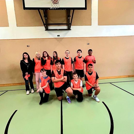 Eine Gruppe junger Menschen in orangefarbenen Westen posiert für ein Foto auf einem Basketballplatz. Sie alle lächeln und machen verschiedene Posen.