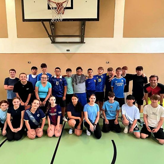 Eine Gruppe junger Leute posiert für ein Foto auf einem Basketballplatz, einige tragen Sporttrikots. Sie lächeln und scheinen Freunde zu sein. Der Platz hat eine grüne Oberfläche.