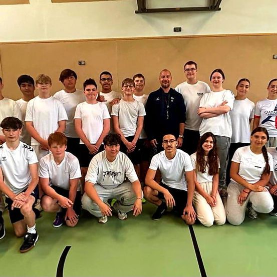 Eine Gruppe von Menschen in weißen T-Shirts und Turnschuhen posiert für ein Foto auf einem Spielfeld. Sie stehen in einer Reihe, einige knien.
