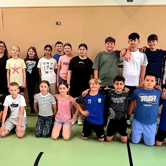 Eine Gruppe von Kindern und einigen Erwachsenen posiert für ein Foto in einer Turnhalle. Einige der Kinder knien, während andere stehen. Sie tragen Freizeitkleidung.