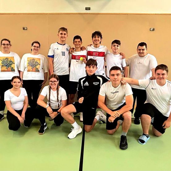 Eine Gruppe junger Athleten posiert für ein Foto in einer Turnhalle, trägt Sporttrikots und Turnschuhe. Einige Leute stehen hinter ihnen.