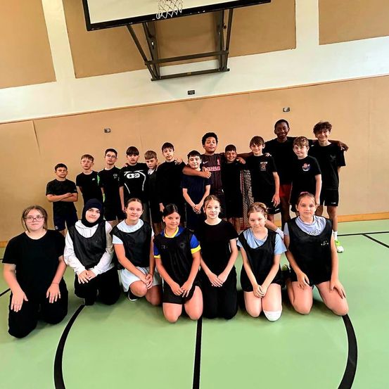 Eine Gruppe junger Menschen, darunter Jungen und Mädchen, posiert für ein Foto auf einem Basketballplatz. Einige tragen Sporttrikots, andere sind in Freizeitkleidung. Im Hintergrund ist ein Basketballkorb zu sehen.