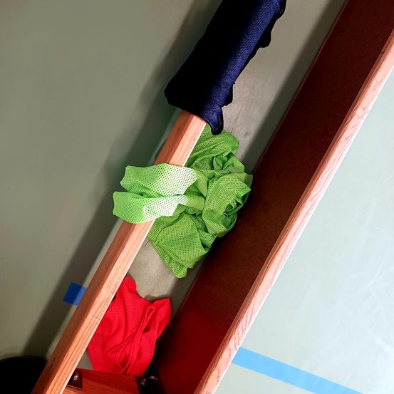 Ein Holzbalken mit einem blauen Tuch oben und einem grünen und roten Tuch darunter. Ein blauer Aufkleber ist an der Wand befestigt.
