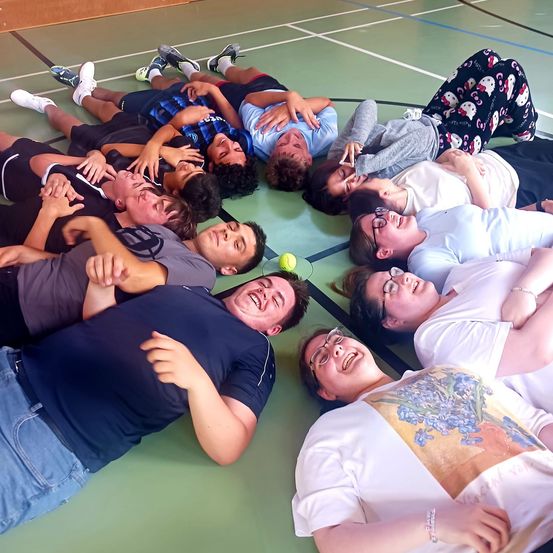 Eine Gruppe von Menschen in lässiger Kleidung liegt in einem Kreis auf einem grünen Boden mit weißen Linien, lacht und lächelt, ein Tennisball ist in der Nähe.