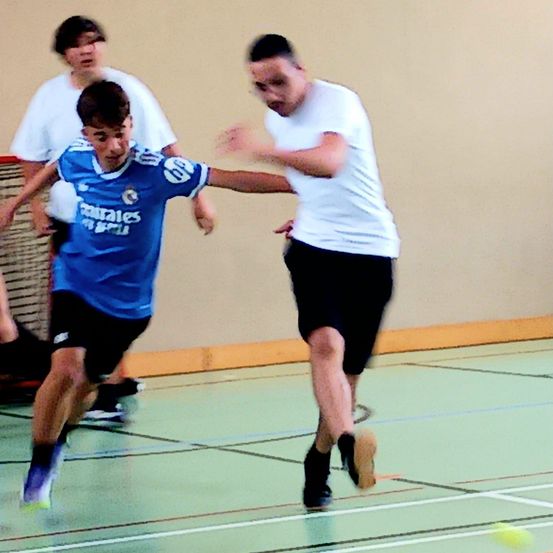 Drei junge Männer spielen Futsal in Innenräumen. Einer in Blau versucht, einem in Weiß auszuweichen. Ein dritter Mann beobachtet im Hintergrund. Der Boden ist mit Linien markiert.