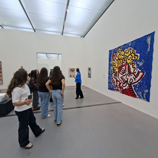 Eine Gruppe von Menschen betrachtet Kunstwerke in einer modernen Galerie. Eine Person untersucht ein großes Stück mit abstrakten Farben und Formen.