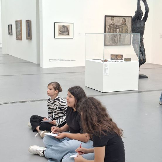Drei junge Mädchen sitzen auf dem Boden in einem Museum. Sie tragen alle schwarze Shirts und schreiben auf Papier. In der Nähe befinden sich gerahmte Gemälde an der Wand und ein Glasvitrine.
