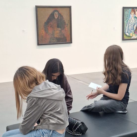 Drei Frauen sitzen auf einer Bank in einem Museum, eine hält einen Stift und Papier. Ein Gemälde einer Frau in Rot und Orange hängt an der Wand hinter ihnen.