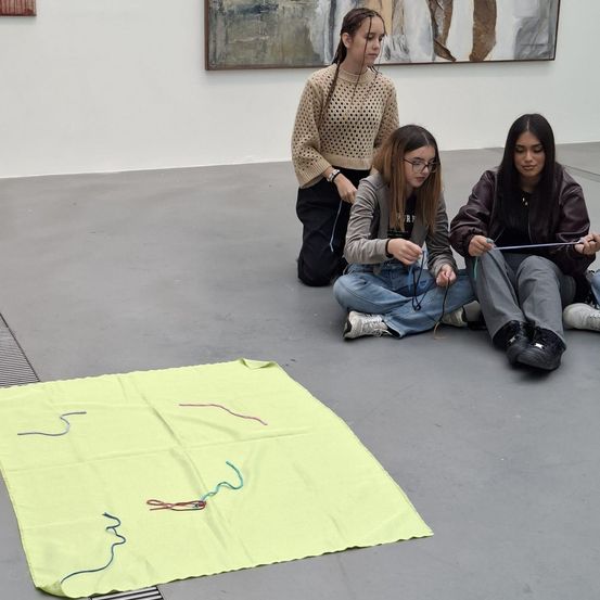Drei junge Frauen sitzen auf dem Boden einer Galerie und beschäftigen sich mit einer kollaborativen Kunstaktivität. Vor ihnen liegt ein gelber Stoff mit farbigen Fäden. An der Wand hinter ihnen befindet sich ein großes, abstraktes Gemälde.