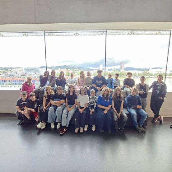 Eine Gruppe von Menschen sitzt auf einer Bank vor einem großen Fenster. Sie lächeln und posieren für ein Foto. Der Raum hat eine Betonwand und -boden.