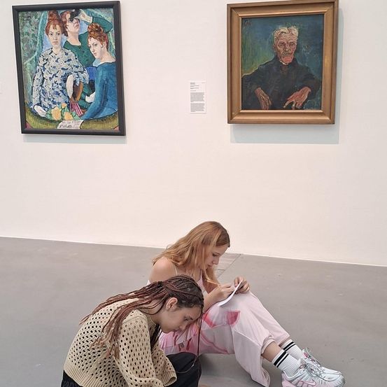 Zwei Frauen sitzen vor Gemälden in einer Kunstgalerie auf dem Boden. Eine Frau schreibt auf Papier.