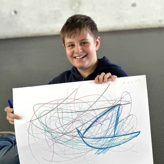 Ein lächelnder Junge hält ein großes Blatt Papier mit farbenfrohen Zeichnungen hoch, einen blauen Stift in der anderen Hand.