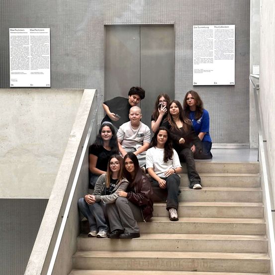 Eine Gruppe junger Menschen sitzt auf einer Treppe in einem Gebäude und posiert für ein Foto. Eine Person macht ein Selfie. An der Wand hinter ihnen befinden sich zwei große Schilder.
