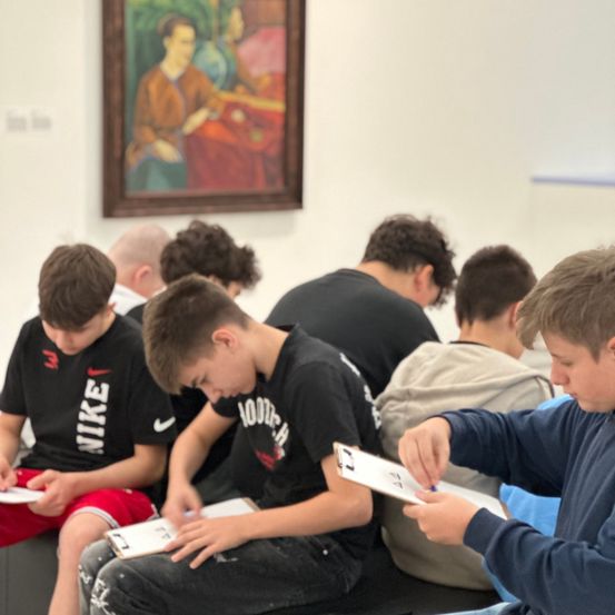 Eine Gruppe von Jungen sitzt in einem Klassenzimmer und schaut auf Papiere. Ein Junge im Nike-Shirt hält einen Marker. Dahinter hängt ein Gemälde an der Wand.