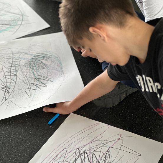 Ein Junge lehnt sich auf einem Tisch vor, konzentriert auf ein Blatt mit Kritzeleien. Ein weiteres Papier mit Kritzeleien liegt vor ihm. Ein blauer Marker liegt auf dem Tisch.