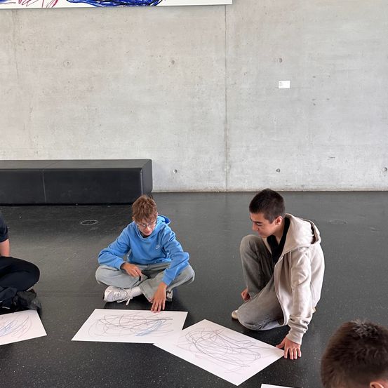 Drei Jungen sitzen auf einem schwarzen Boden, einer von ihnen schaut auf ein Blatt Papier mit einer Spirale darauf. Hinter ihnen ist eine Betonwand mit einem großen Kunstwerk obenauf.