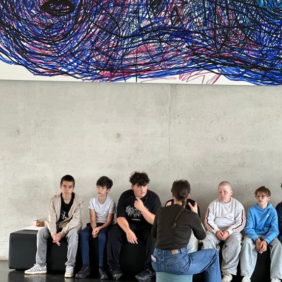 Eine Gruppe von Jungen und eine Frau sitzen auf einer Bank unter einem großen abstrakten Gemälde an einer weißen Wand. Die Frau, die in der Mitte sitzt, scheint die Jungen zu unterrichten oder zu interviewen.
