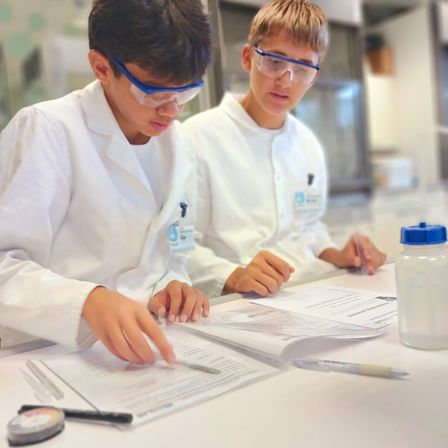 Zwei Schüler in Laborkitteln und Schutzbrillen arbeiten gemeinsam an einem Labortisch und untersuchen Papiere und verwenden eine Pipette.