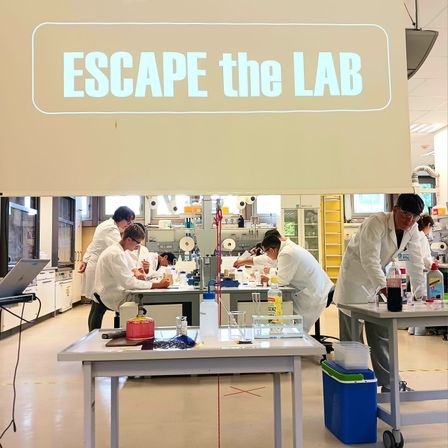 Mehrere Studenten in Laborkittel arbeiten an Experimenten in einem Labor. Ein Banner mit den Worten 'ESCAPE the LAB' hängt über ihnen.