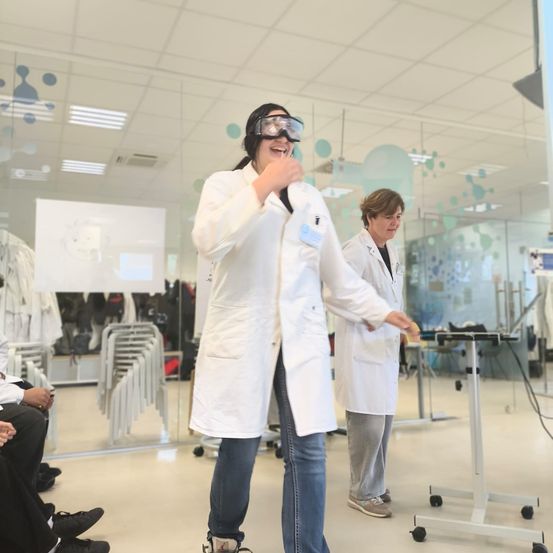 Zwei Frauen in weißen Laborkittel und Schutzbrillen sind in einem Glasraum. Eine steht und lächelt, die andere sieht sie an. Dahinter steht eine Tafel und Stühle.