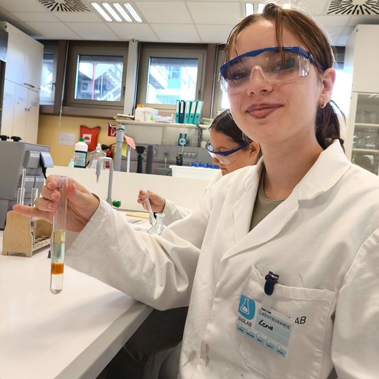 Eine Frau in einem Laborkittel hält ein Reagenzglas mit einer Flüssigkeitsprobe und trägt Schutzbrillen. Hinter ihr steht eine weitere Person in einem Laborkittel, die in einem Labor mit verschiedenen Geräten arbeitet.