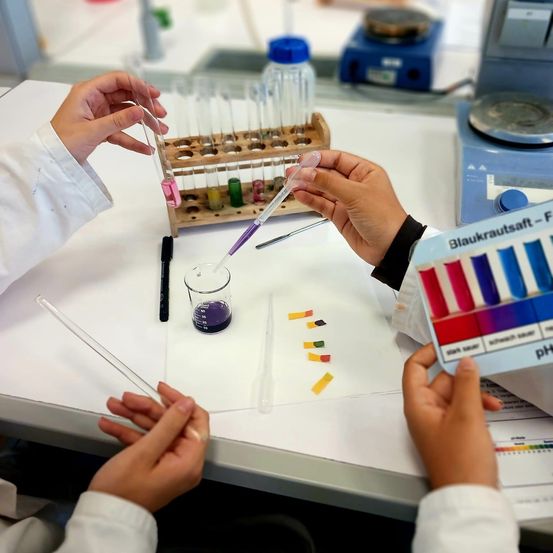 Laboranten mit Laborkittel führen ein Chemieexperiment durch. Sie verwenden eine Pipette, um Flüssigkeit in ein kleines Teströhrchen zu übertragen. Verschiedene Laborgeräte stehen auf dem Tisch.
