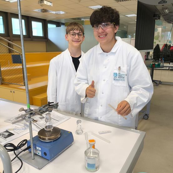 Zwei Schüler in weißen Laborkitteln stehen in einem Labor, einer hält einen Stock, lächelt und posiert für ein Foto. Ausrüstung und ein Tisch mit einer Glasflasche sind im Hintergrund zu sehen.