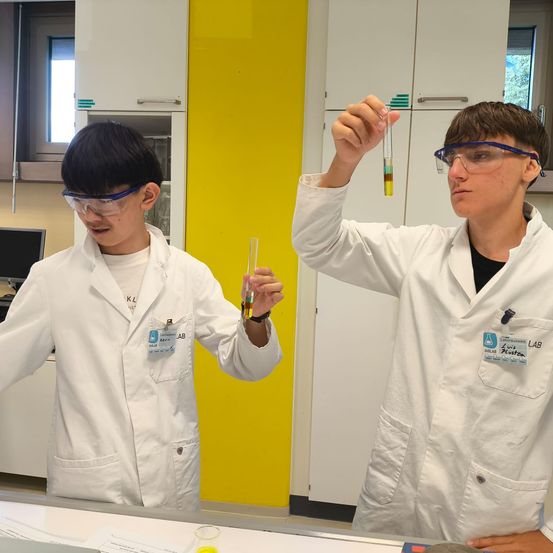 Zwei junge Personen in weißen Laborkittel und Schutzbrillen führen ein wissenschaftliches Experiment in einem Labor durch. Einer hält ein Reagenzglas mit einer Flüssigkeit, die in verschiedenen Farben geschichtet ist.