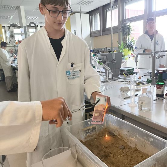 In einem Labor hält ein Schüler eine brennende Banknote in einem Sandbehälter. Zwei weitere Schüler in Laborkittel beobachten. Das Labor hat einen Schreibtisch, Flaschen und eine Pflanze.