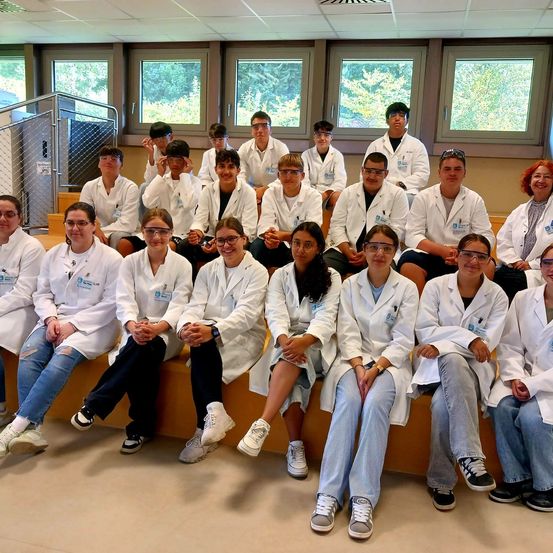 Eine Gruppe von Studenten in weißen Laborkittel und Brillen posiert für ein Foto in einem Klassenzimmer mit großen Fenstern.