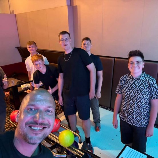 Eine Gruppe junger Männer steht in einer Bowlingbahn, lächelt und posiert für ein Foto. Ein Mann hält einen Bowlingball.