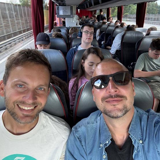 Zwei Männer lächeln und posieren für ein Foto in einem Bus mit anderen Passagieren.