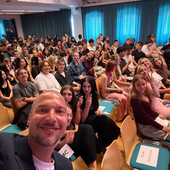 Eine große Gruppe von Menschen sitzt in einem Raum, wahrscheinlich einem Hörsaal, viele lächeln und einige halten Papiere. Ein Mann vorne macht ein Selfie.