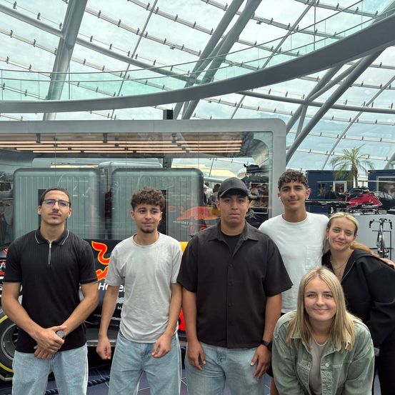 Fünf Personen stehen vor einem roten Auto, wahrscheinlich bei einer Veranstaltung. Sie alle lächeln und scheinen für ein Foto zu posieren.