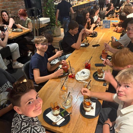 Eine Gruppe junger Leute sitzt an einem langen Tisch in einem Restaurant, jeder mit einem Glas Getränk und einem Dessert vor sich.