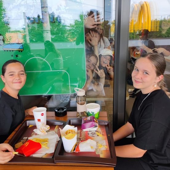 Zwei Mädchen sitzen an einem McDonald's-Tisch mit Essenstabletts und lächeln und posieren für ein Foto mit anderen im Hintergrund.