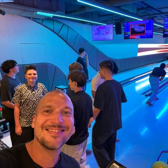 Eine Gruppe von Menschen ist in einer Bowlingbahn, wobei ein Mann ein Selfie macht, während andere ein Bowlingspiel auf der Bahn beobachten.