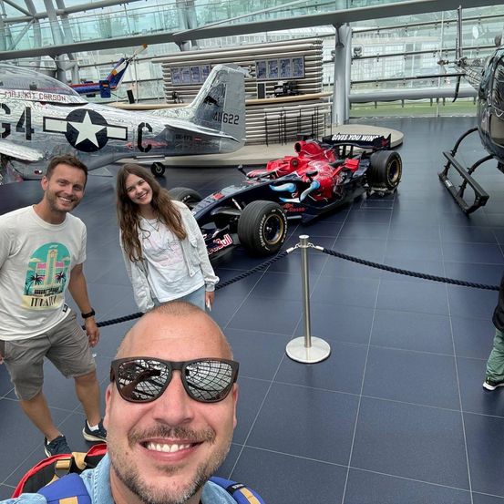 Drei Personen stehen vor einem roten Rennwagen und einem silbernen Flugzeug in einem Museum. Der Mann links trägt ein weißes T-Shirt mit grafischem Design.
