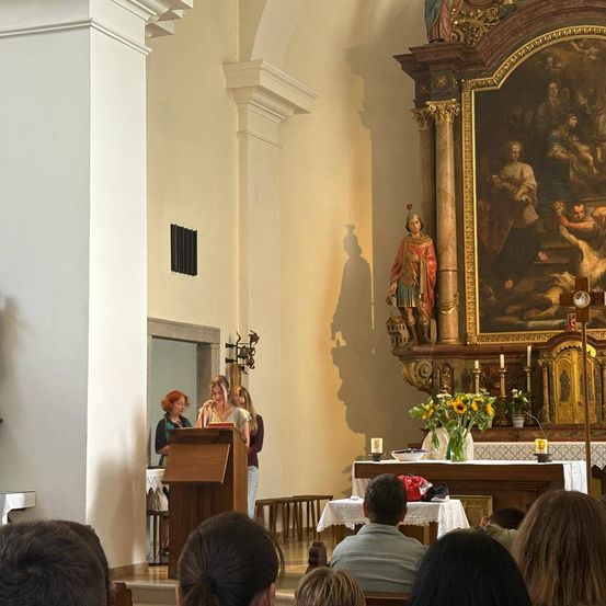 Eine Gruppe von Menschen versammelt sich in einer Kirche, wobei eine Frau an einem Podium steht und andere auf den Kirchenbänken sitzen. Ein großes Gemälde schmückt die Wand hinter dem Altar.
