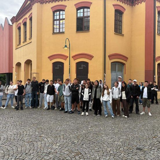 Eine Gruppe von Menschen, wahrscheinlich Schüler, steht vor einem gelben Gebäude mit Bogenfenstern und einer Straßenlaterne. Sie sind lässig gekleidet und scheinen für ein Foto zu posieren.