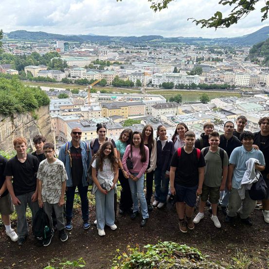 Eine Gruppe junger Menschen und Erwachsener steht auf einem Felsen mit Blick auf eine Stadt mit vielen Gebäuden und einem Fluss.