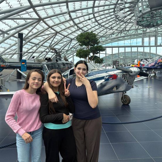 Drei Frauen posieren für ein Foto vor einem Flugzeug in einem Museum. Die Frau links trägt einen rosa Pullover, die mittlere ein schwarzes Oberteil und die rechte ein blaues Shirt. Hinter ihnen steht ein silbernes Flugzeug mit Propeller.