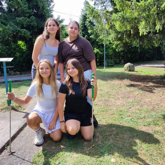 Vier junge Frauen posieren für ein Foto auf einem Golfplatz. Zwei knien mit Golfschlägern, und zwei stehen hinter ihnen. Bäume und ein kleiner Stein sind im Hintergrund.