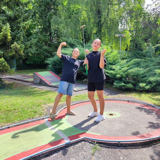 Zwei junge Mädchen posieren für ein Foto auf einem Minigolfplatz, halten Golfschläger und lächeln. Der Platz ist von grünen Büschen und Bäumen umgeben.