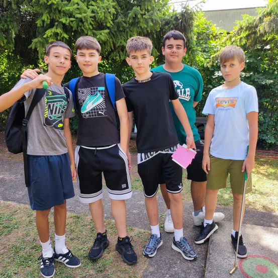 Fünf Jungen stehen auf einem Betonweg und lächeln für ein Foto. Sie tragen Freizeitkleidung und Turnschuhe, einer hält einen Golfschläger. Hinter ihnen bilden Grünflächen und Bäume eine natürliche Kulisse.