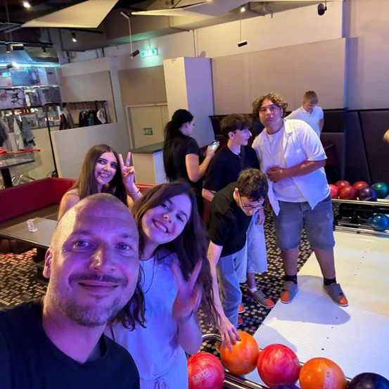 Eine Gruppe von Menschen lächelt und posiert für ein Foto in einer Bowlingbahn, mit Bowlingkugeln und Pins im Hintergrund.