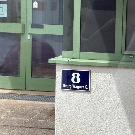 Ein blaues Schild mit der Nummer 8 und dem Namen Georg-Wagner-G. an einer weißen Wand. Das Schild befindet sich neben einer grünen Tür mit Glasscheiben.