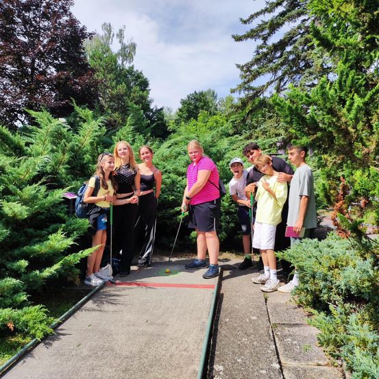 Eine Gruppe von Menschen, darunter junge Mädchen und Jungen, stehen auf einem Betonweg in einem Park, halten Golfschläger und posieren für ein Foto. Sie sind von grünen Bäumen und Büschen umgeben.