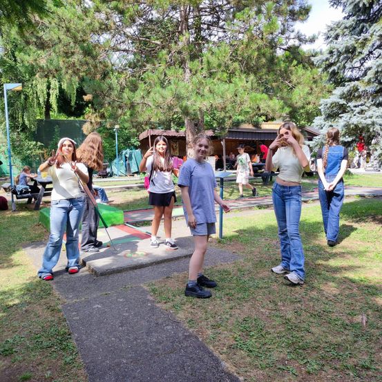 Eine Gruppe junger Frauen steht in einem Park, eine von ihnen macht ein Friedenszeichen. Hinter ihnen befindet sich ein Minigolfplatz mit einem kleinen Gebäude.