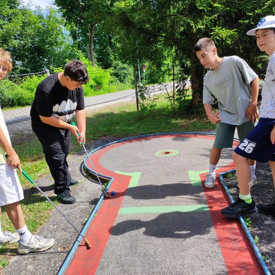 Vier Jungen spielen an einem sonnigen Tag Minigolf. Ein Junge ist dabei, den Ball zu schlagen. Die anderen schauen zu.